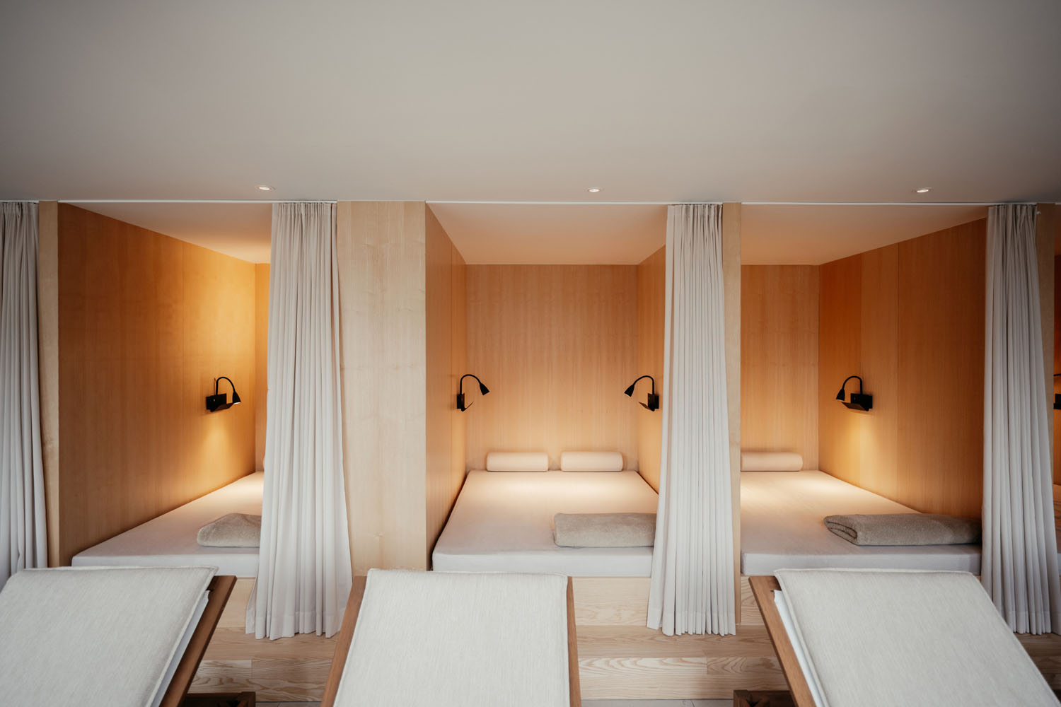 Drei gemütliche Nischen aus Holz sind jeweils mit einem Bett mit weißer Bettwäsche, Kissen, an der Wand befestigten Leselampen und neutralen Vorhängen für mehr Privatsphäre ausgestattet; im Vordergrund stehen zwei gepolsterte Liegestühle.