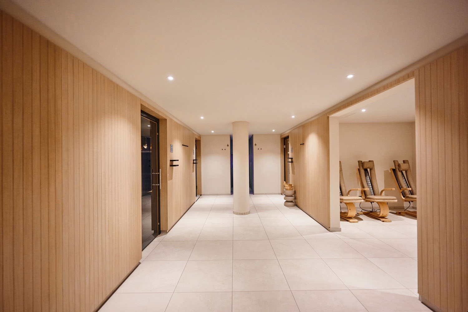 Moderner Spa-Flur mit hellen Holzvertäfelungen, weißem Fliesenboden und Einbauleuchten. Auf der rechten Seite sind in einem Entspannungsbereich Liegestühle aus Holz aufgestellt.