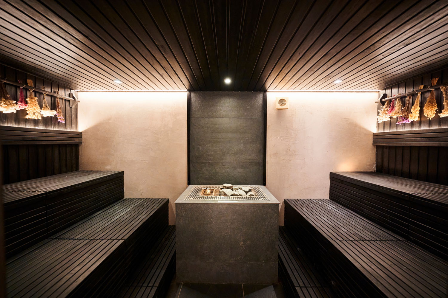 Moderne Sauna mit dunklen Holzbänken, einem zentralen Steinofen mit Steinen darauf und getrockneten Kräutern, die unter warmer Stimmungsbeleuchtung an den Wänden hängen.
