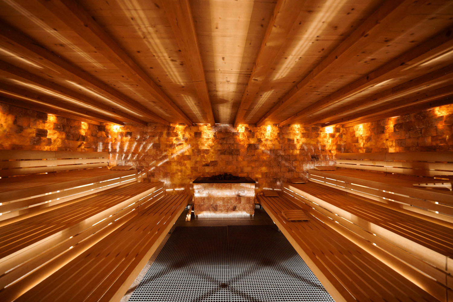 Innenansicht einer modernen Sauna mit Holzbänken auf beiden Seiten und warmer, stimmungsvoller Beleuchtung, die von einer Steinwand am Ende des Raums reflektiert wird. Das Foto wurde aus einem niedrigen Blickwinkel aufgenommen, wodurch die Decke und die Symmetrie besonders hervorgehoben werden.