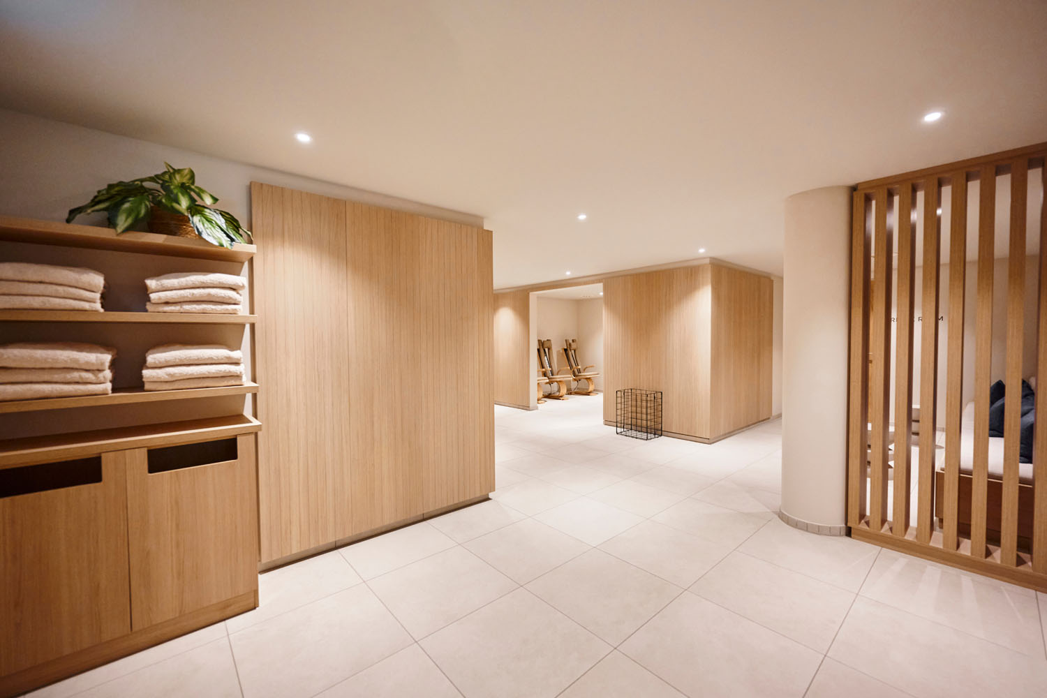 Modernes Spa-Interieur mit Holzregalen, auf denen ordentlich gefaltete Handtücher liegen, hellen Holzvertäfelungen, einem beigen Fliesenboden und sanften Deckenleuchten, die eine ruhige, minimalistische Atmosphäre schaffen.