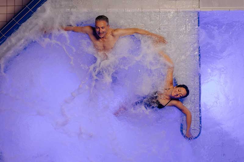 Zwei Personen, ein Mann und eine Frau, die sich in einem sprudelnden Whirlpool oder Pool mit blauer Beleuchtung entspannen, von oben betrachtet. Beide scheinen das Wasser zu genießen und lehnen sich an den Rand.