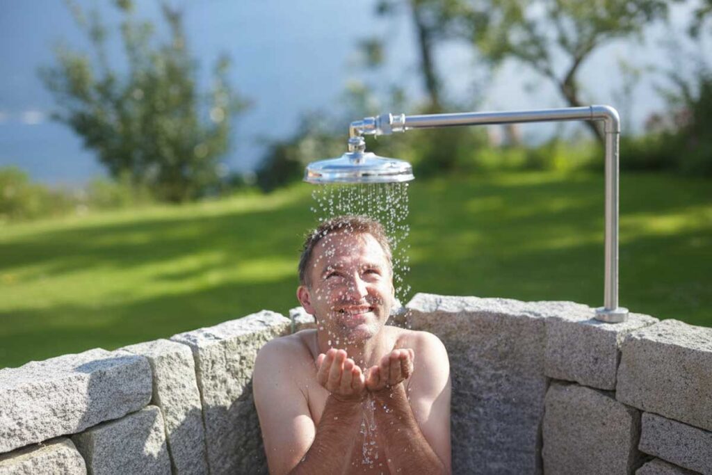 Ein Mann steht unter einer Außendusche, fängt das Wasser mit den Händen auf und lächelt. Die Dusche ist von einer niedrigen Steinmauer umgeben, im Hintergrund sind Gras und Bäume zu sehen.