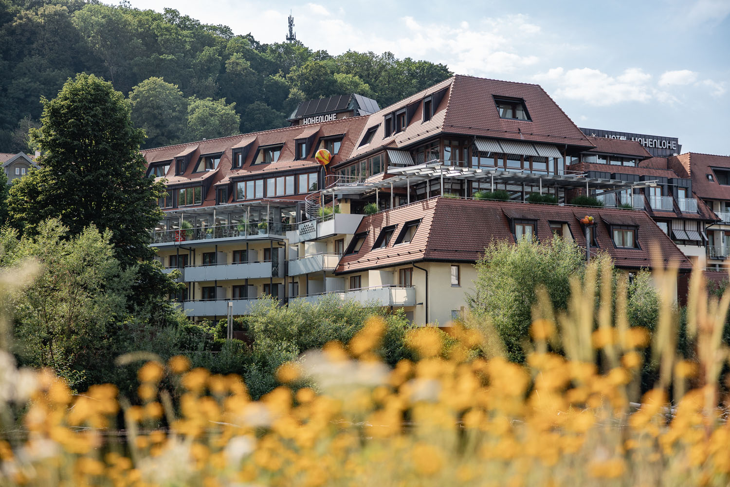 Am Fuße eines bewaldeten Hügels steht ein großes, modernes Hotel mit roten Dächern und Balkonen. Im Vordergrund blühen gelbe Wildblumen, und der Himmel ist teilweise bewölkt.