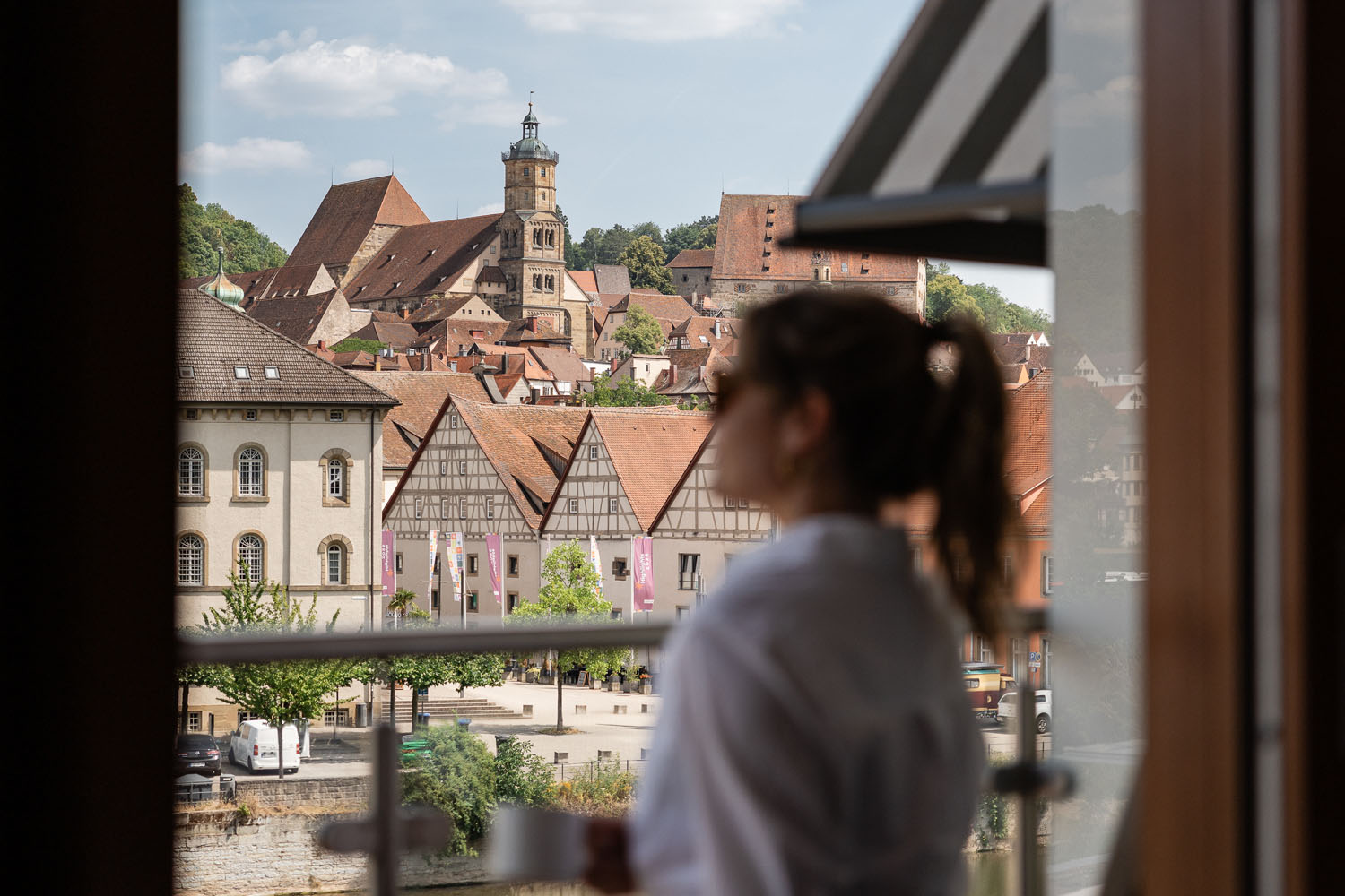 Eine Frau in einem weißen Gewand steht auf einem Balkon, hält eine Tasse in der Hand und blickt auf eine malerische europäische Stadt mit Fachwerkhäusern und einem markanten Kirchturm im Hintergrund.