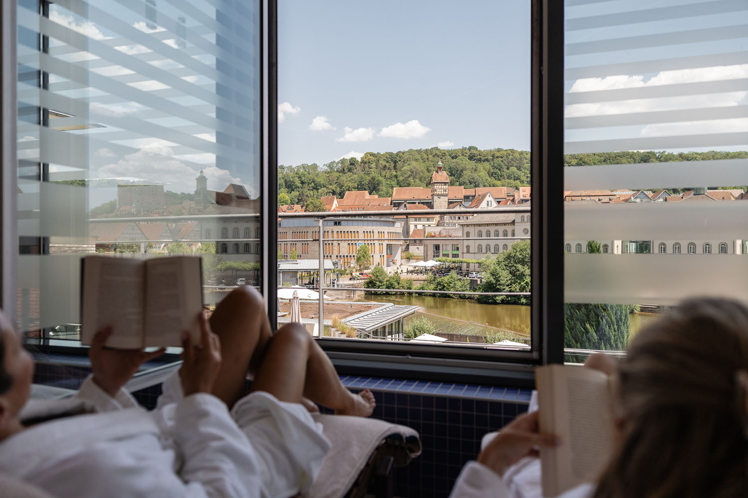 Zwei Menschen in weißen Gewändern entspannen sich und lesen an einem großen Fenster mit malerischem Blick auf einen Fluss, historische Gebäude und grüne Hügel unter einem teilweise bewölkten Himmel.