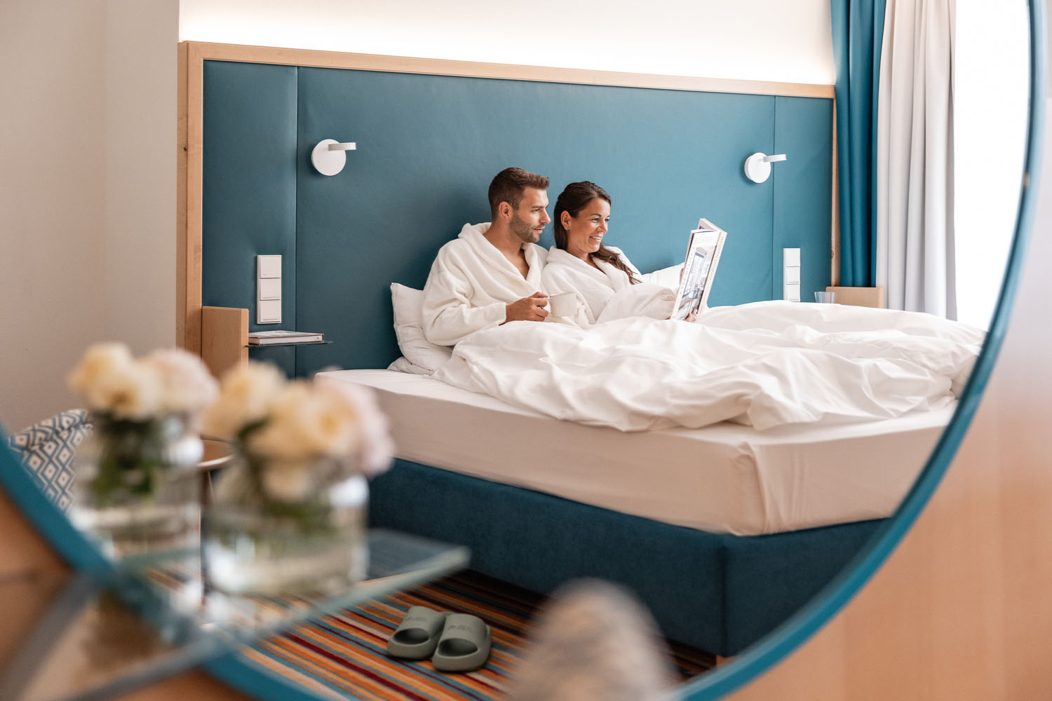 Ein Paar in weißen Gewändern sitzt auf einem Bett, lächelt und liest in einem modernen Hotelzimmer eine Zeitschrift. Frische Blumen und Hausschuhe sind zu sehen, und die Szene spiegelt sich in einem runden Spiegel wider.