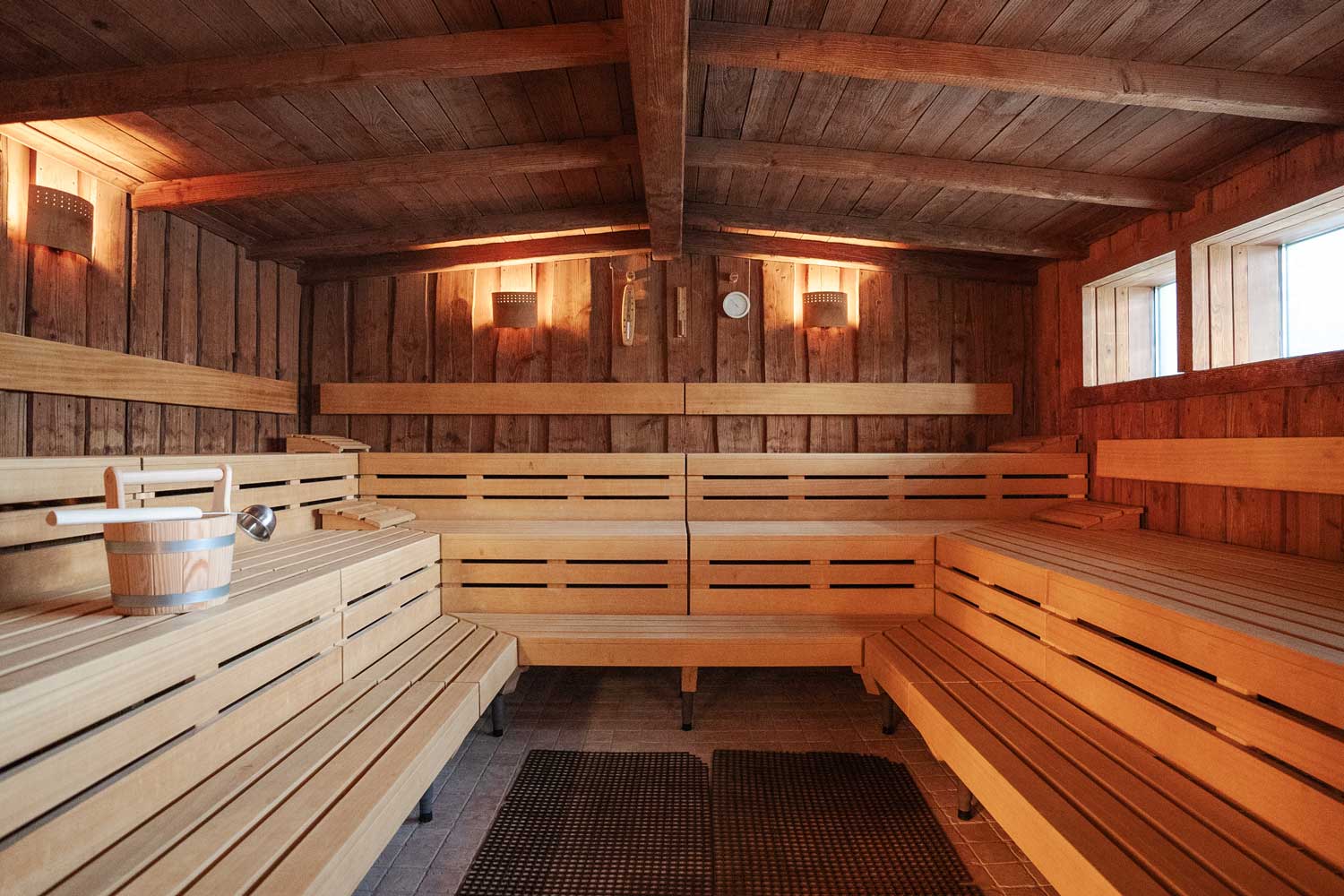 Innenraum einer Holzsauna mit Sitzbänken entlang der Wände, einem Eimer mit einer Schöpfkelle auf einer der Bänke, sanfter Beleuchtung an den Holzwänden und zwei Fenstern, durch die Tageslicht hereinfällt.