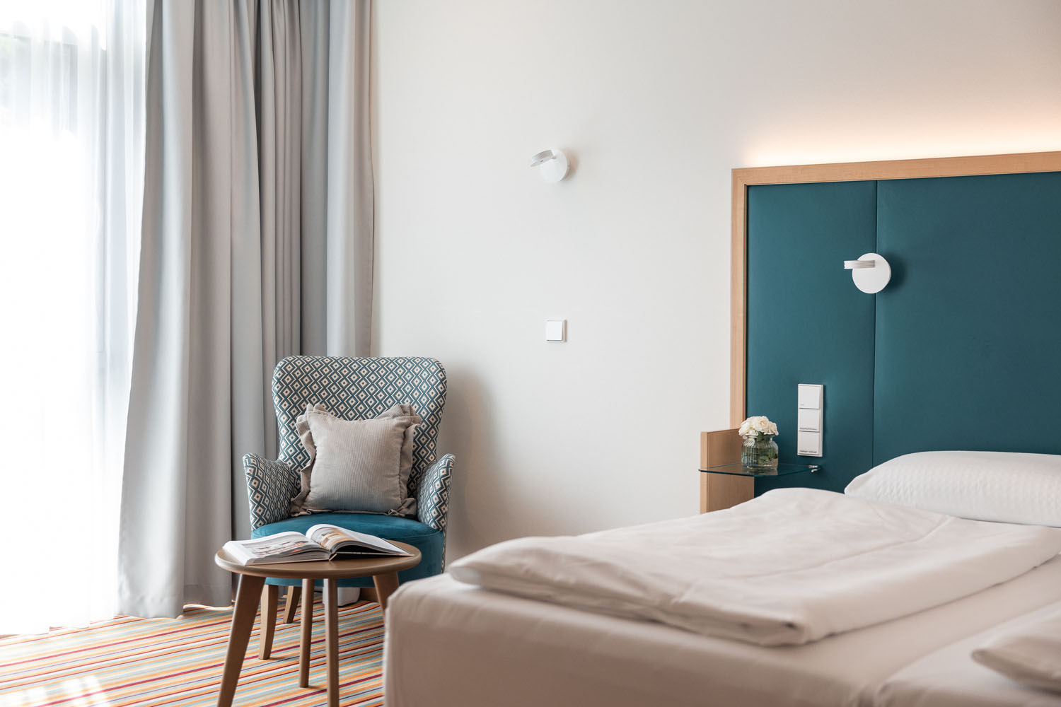 Ein modernes Hotelzimmer mit einem ordentlich gemachten Bett, einem blaugrünen Kopfteil, einem gemusterten Sessel mit einem Kissen, einem kleinen Tisch, auf dem ein aufgeschlagenes Buch liegt, und einer Vase mit Blumen an der Wand. Durch die transparenten Vorhänge fällt Licht herein.