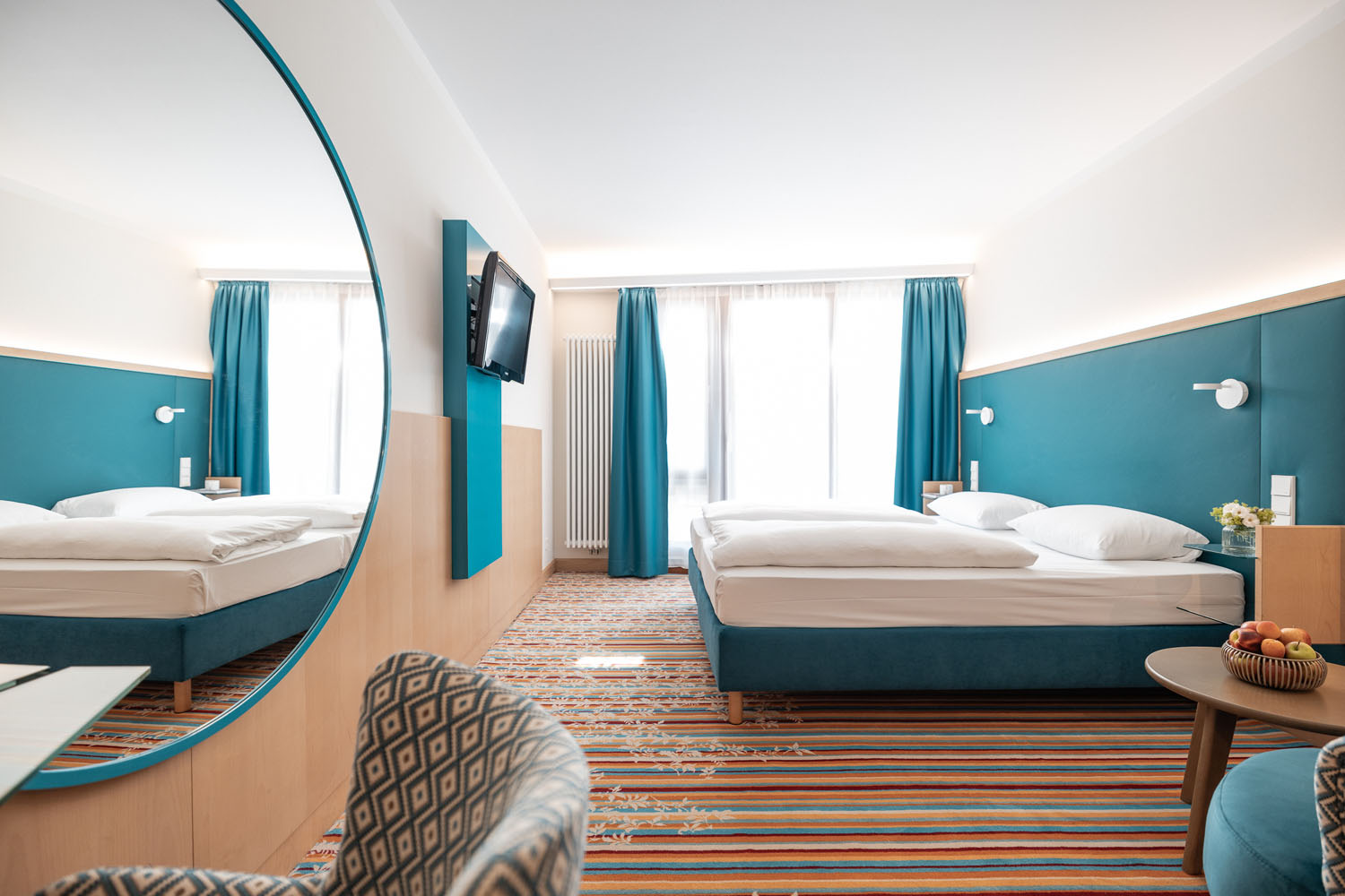 Modernes Hotelzimmer mit blau-weißer Einrichtung, einem Doppelbett, einem gestreiften Teppich, einem an der Wand montierten Fernseher, einem großen runden Spiegel, einem Sessel und einem kleinen Tisch mit einem Obstkorb, alles gut beleuchtet durch das natürliche Licht, das durch das Fenster fällt.