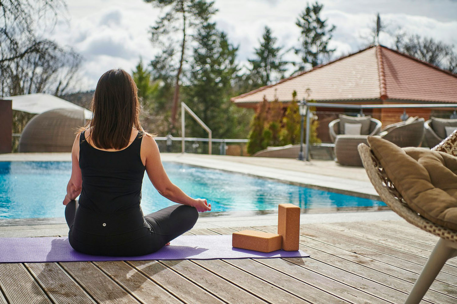 Eine Frau sitzt mit gekreuzten Beinen auf einer Yogamatte am Pool und meditiert im Freien. In der Nähe liegen Yogablöcke. Im Hintergrund sind unter einem bewölkten Himmel Bäume und Terrassenmöbel zu sehen.