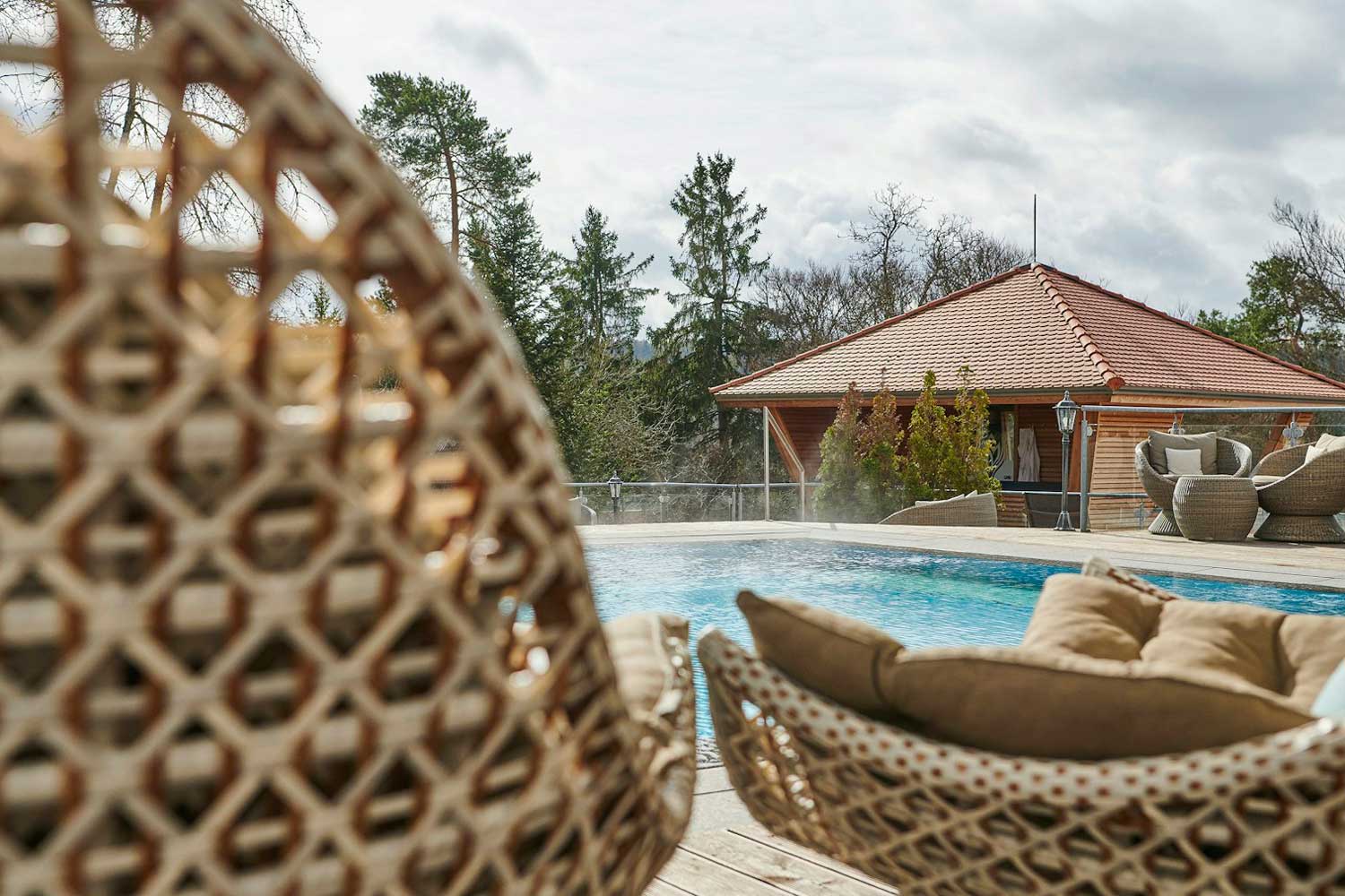 Szene am Pool mit geflochtenen Liegestühlen im Vordergrund, einem klaren Schwimmbecken und einem Holzgebäude mit rotem Ziegeldach im Hintergrund, umgeben von Bäumen unter einem bewölkten Himmel.