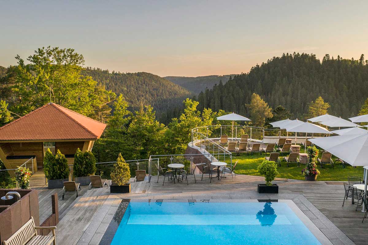 Ein ruhiger Außenpool mit Liegestühlen und Sonnenschirmen bietet bei Sonnenuntergang unter klarem Himmel einen Blick auf einen üppig grünen Wald und die fernen Hügel.