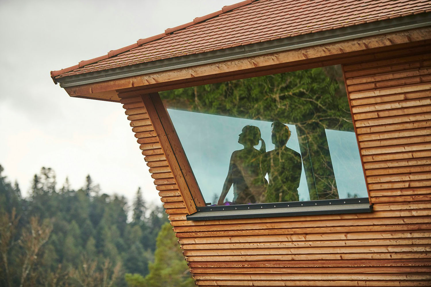 Durch ein großes Fenster eines modernen Holzgebäudes sind zwei Personen als Silhouetten zu sehen; draußen sind Bäume und ein bewölkter Himmel zu erkennen.