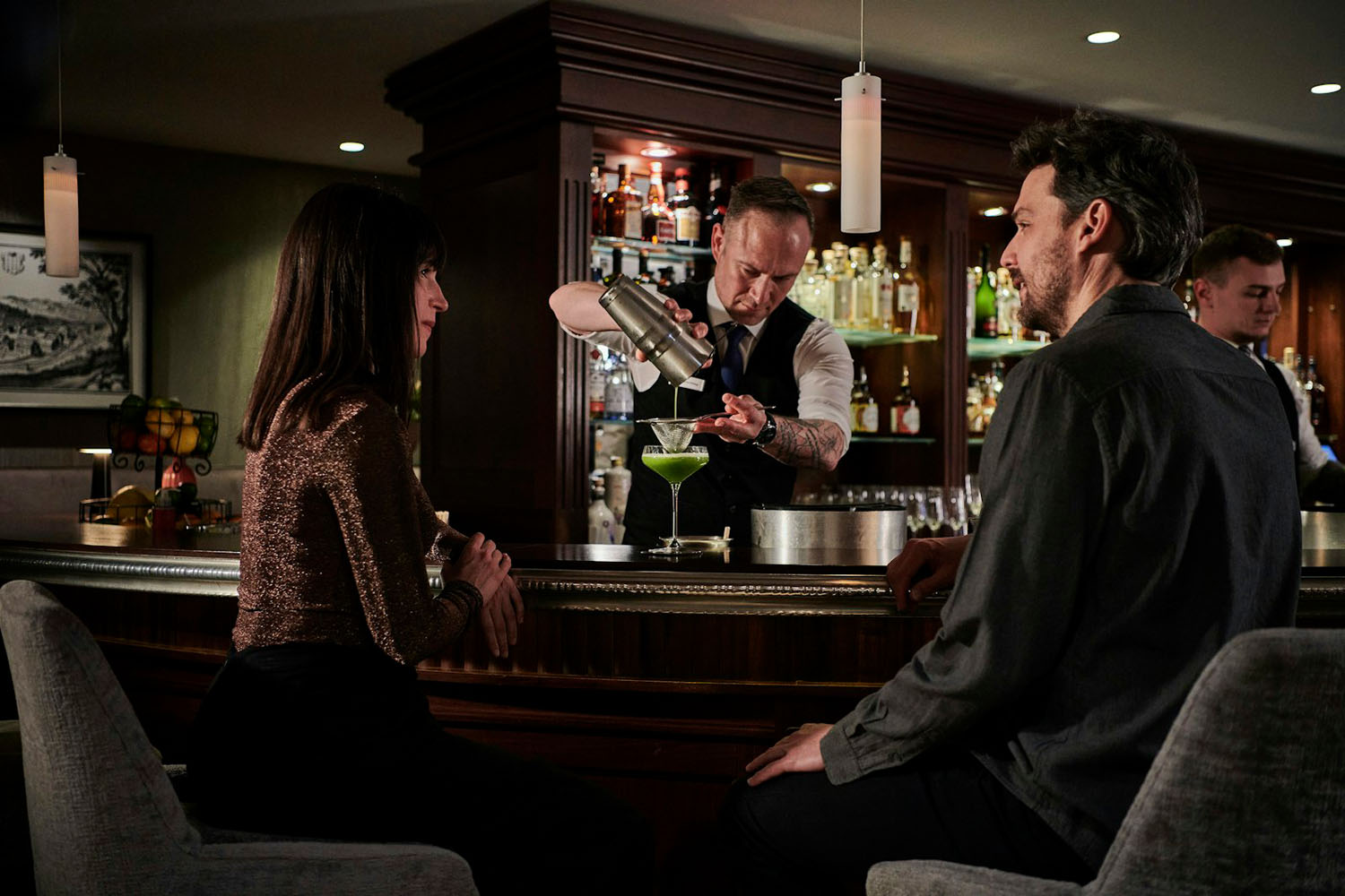 Ein Barkeeper schenkt einen grünen Cocktail in ein Glas ein, während ein Mann und eine Frau an einer schummrig beleuchteten Bar sitzen und sich unterhalten. Im Hintergrund sind Regale mit Flaschen und eine Obstschale zu sehen.
