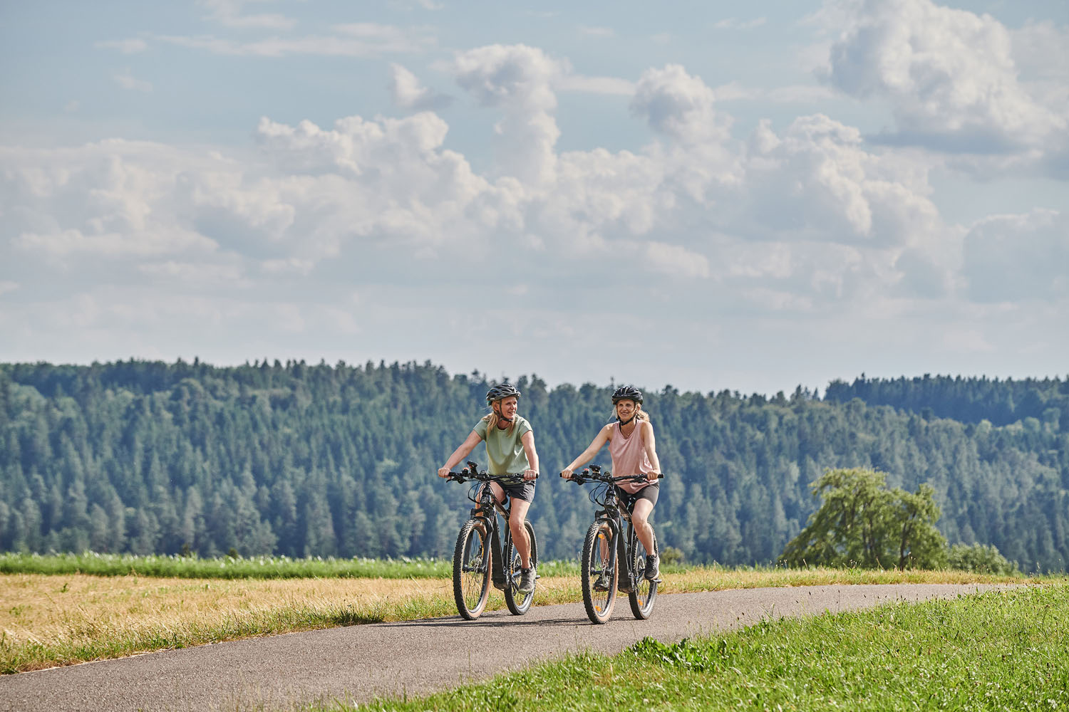 Zwei Personen fahren mit dem Fahrrad auf einem asphaltierten Weg durch grasbewachsene Felder und tragen dabei Helme. Im Hintergrund sind ein Wald und ein blauer Himmel mit vereinzelten Wolken zu sehen.