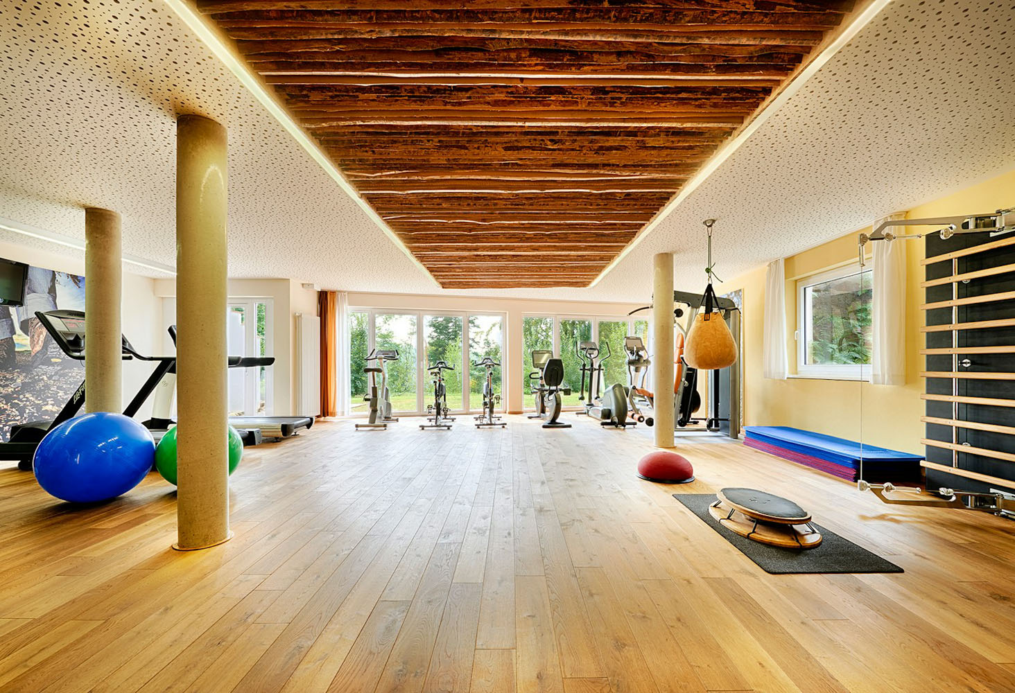 Ein heller Fitnessraum mit Holzboden, Heimtrainern, Laufbändern, einem Boxsack, Gymnastikbällen, Matten und verschiedenen Fitnessgeräten. Große Fenster lassen Tageslicht herein.