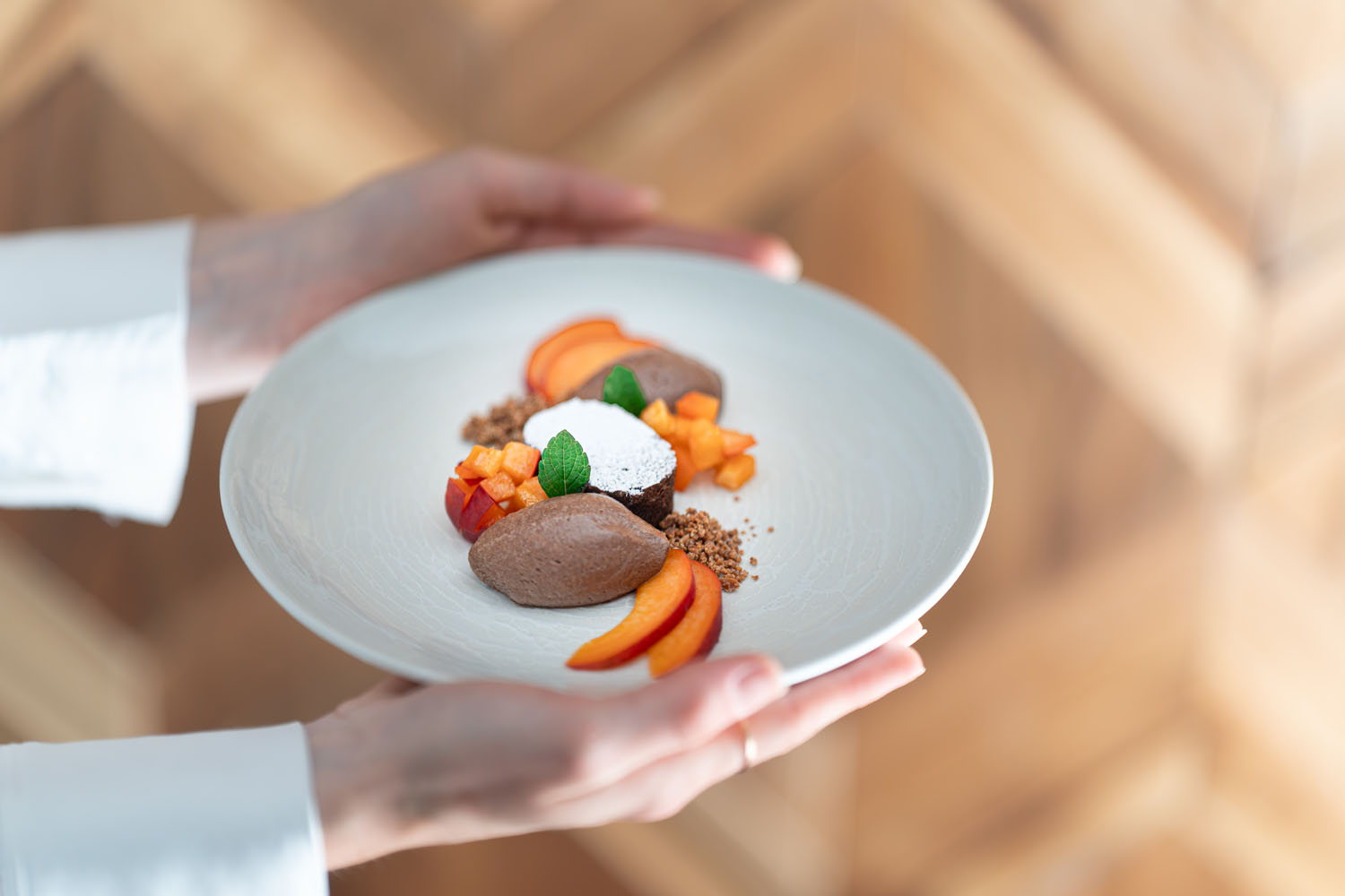 Vor einem unscharfen Holzhintergrund hält eine Person einen weißen Teller mit Schokoladenmousse, Obstwürfeln, Müsli, Minzblättern und Pfirsichscheiben in der Hand.