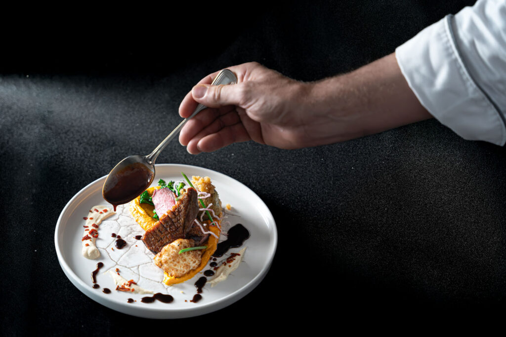 Die Hand eines Kochs träufelt mit einem Löffel eine dunkle Soße über ein kunstvoll angerichtetes Gourmetgericht auf einem weißen Teller, auf dem bunte Gemüsesorten und Beilagen vor schwarzem Hintergrund angerichtet sind.