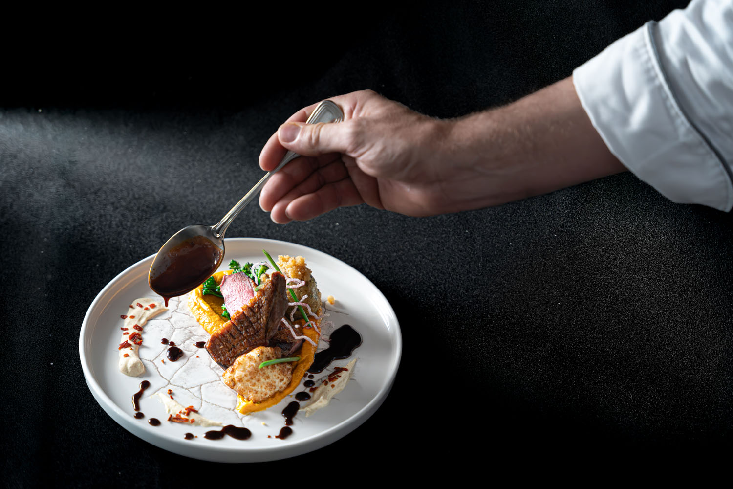 Die Hand eines Kochs träufelt mit einem Löffel eine dunkle Soße über ein kunstvoll angerichtetes Gourmetgericht auf einem weißen Teller, auf dem bunte Gemüsesorten und Beilagen vor schwarzem Hintergrund angerichtet sind.