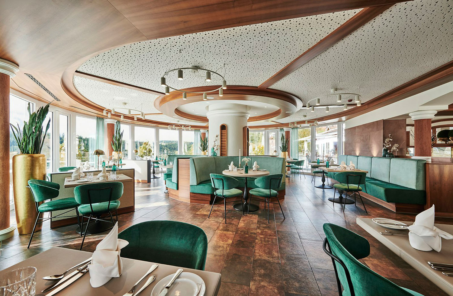 Modernes Restaurantinterieur mit grünen Samtstühlen, runden Holzdekorationen an der Decke und großen Fenstern, durch die Tageslicht hereinströmt. Die Tische sind mit gefalteten Servietten und Gläsern gedeckt, was eine elegante Atmosphäre beim Essen schafft.