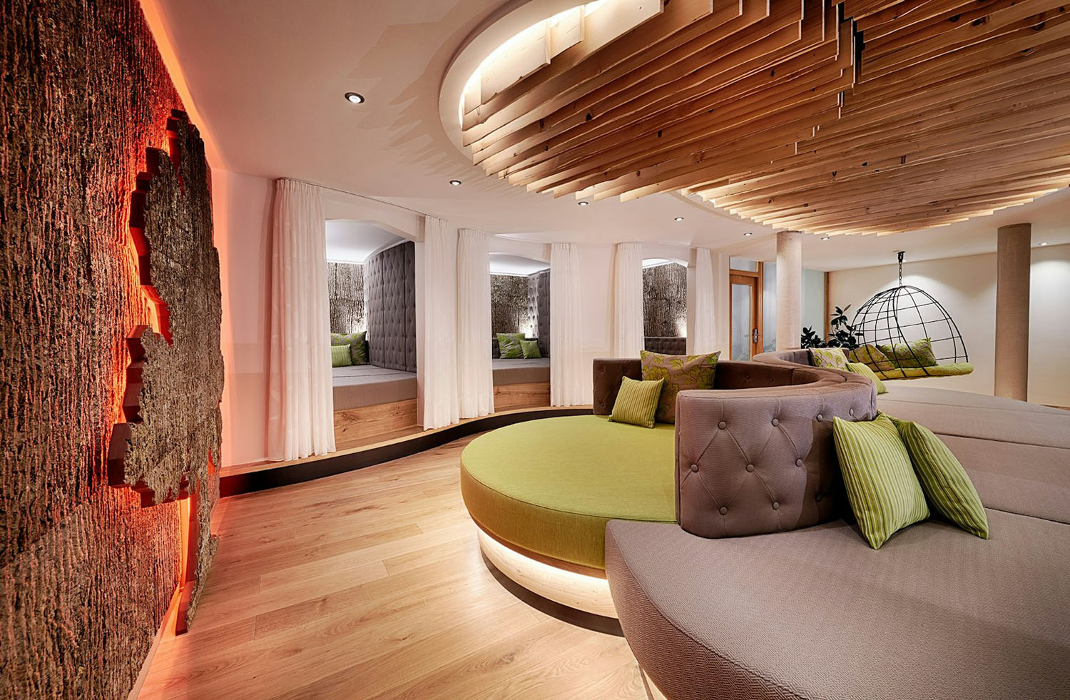 Moderne Spa-Lounge mit geschwungenen grünen Sofas, Holzböden, einer strukturierten Wand mit roter Hintergrundbeleuchtung, Holzpaneelen an der Decke und gemütlichen, mit Vorhängen versehenen Nischen mit Betten. Im Hintergrund sind ein Hängesessel und Pflanzen zu sehen.