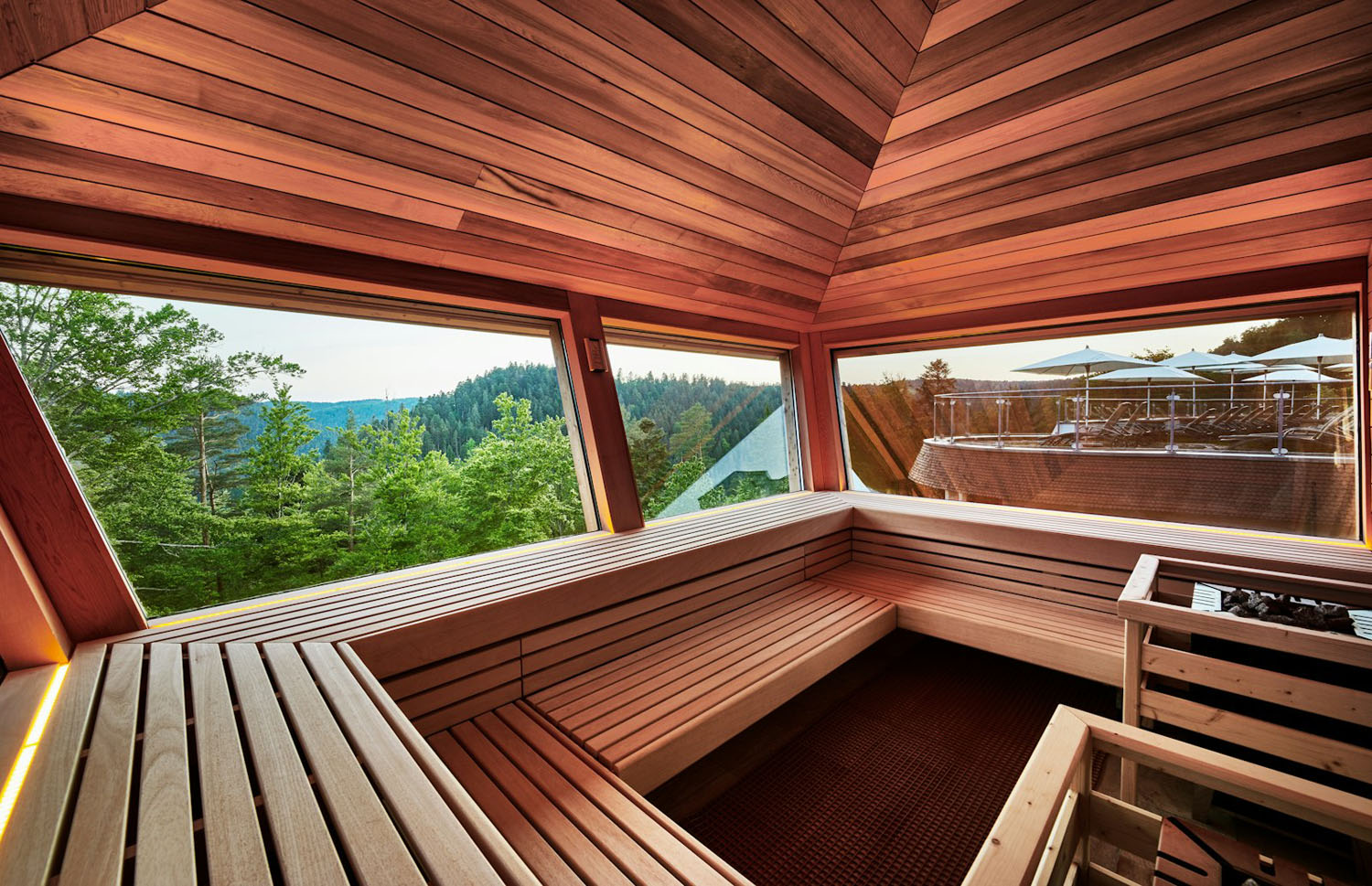 Eine moderne Holzsauna mit großen Fenstern bietet einen malerischen Blick auf grüne Bäume und ferne Hügel. Auf der Terrasse draußen sind Sitzgelegenheiten mit Sonnenschirmen zu sehen. Warmes Sonnenlicht strömt durch die Fenster.