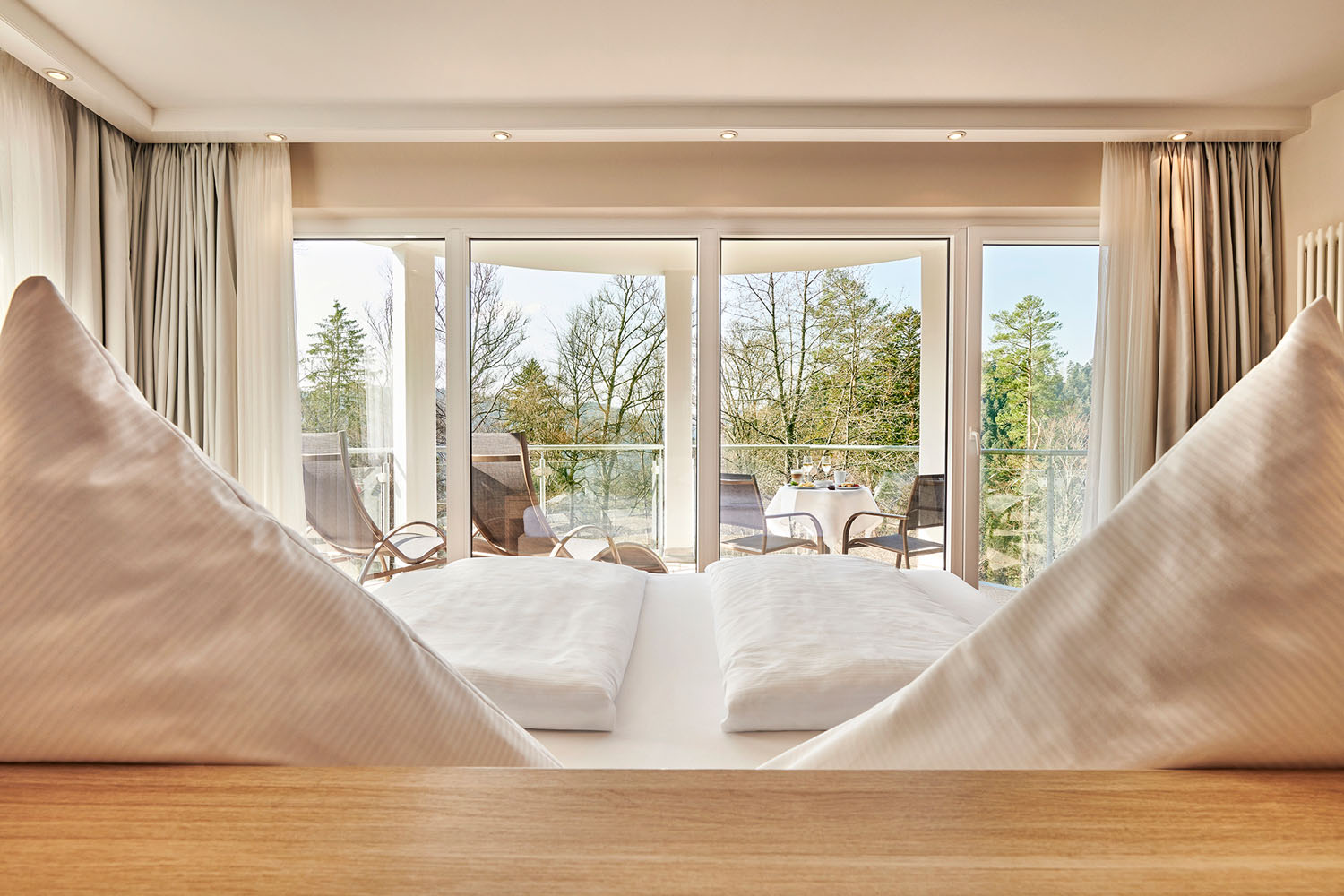 Ein helles Hotelzimmer mit zwei ordentlich gemachten Betten, großen Fenstern und einem Balkon mit Stühlen und einem Tisch, von dem aus man auf die Bäume draußen blickt. Natürliches Licht durchflutet den Raum und schafft eine gemütliche und einladende Atmosphäre.