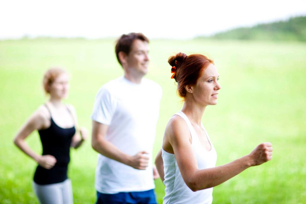 Drei Personen – zwei Frauen und ein Mann – gehen spazieren oder treiben Sport im Freien auf grünem Rasen. Sie tragen Sportkleidung und scheinen die Aktivität in einer hellen, natürlichen Umgebung zu genießen.