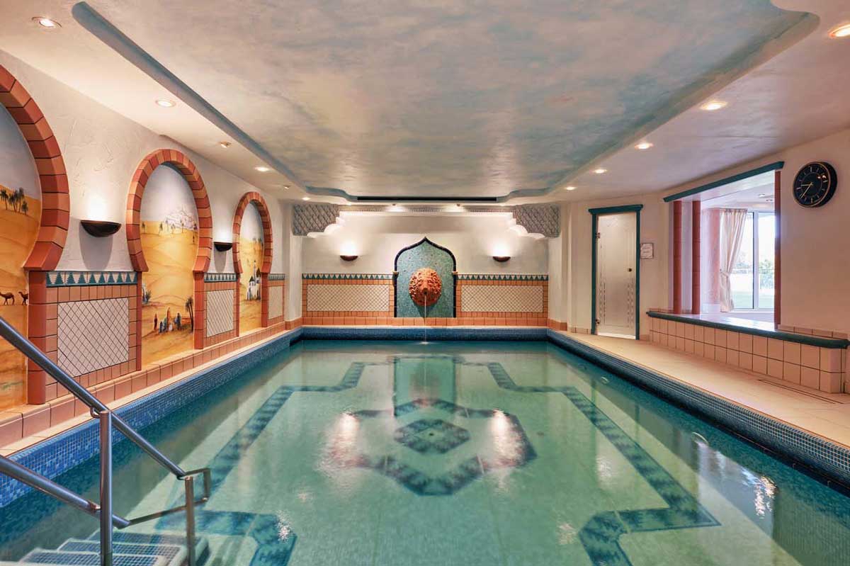 Hallenbad mit einem Fliesenmuster auf dem Boden, umgeben von Wandbögen mit Wüstenmotiven und einem zentralen Wasserspiel in Form eines Löwenkopfes.