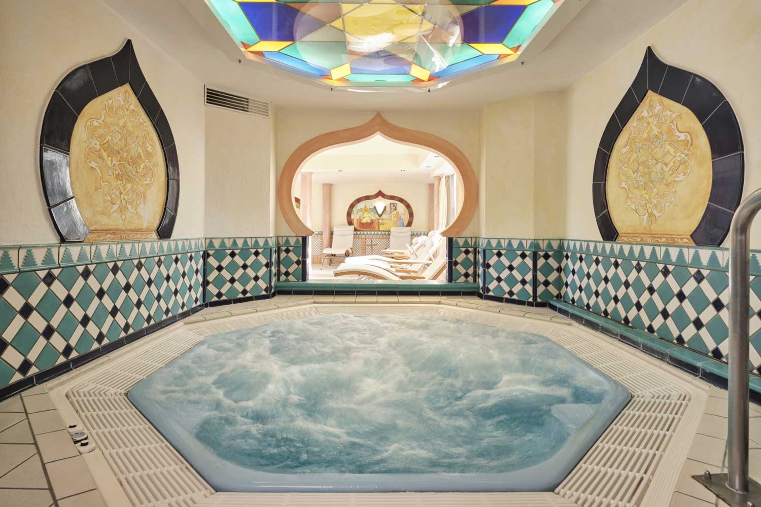 Ein Innen-Whirlpool in einem Raum mit Bögen im marokkanischen Stil, Buntglasfenstern und blau-weiß gefliesten Wänden.
