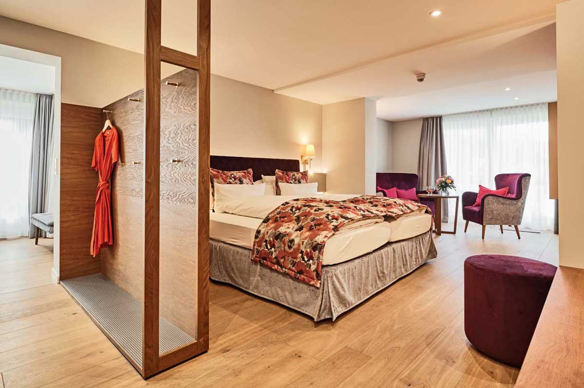 Hotelzimmer mit einem Kingsize-Bett, einem Sichtschutz aus Holz, an dem ein Bademantel hängt, und einer Sitzecke mit violetten Stühlen.