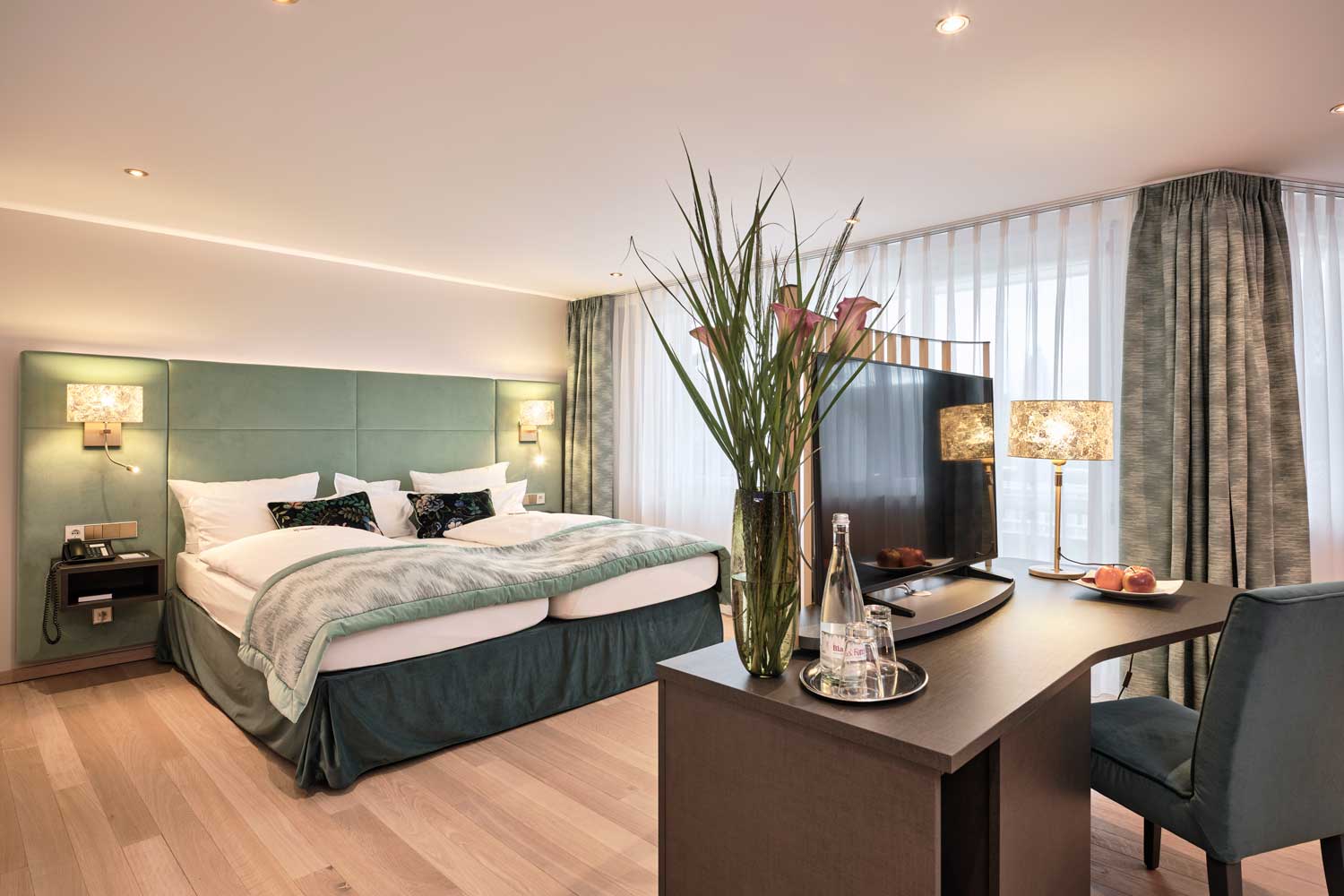 Hotelzimmer mit einem Kingsize-Bett, einem Schreibtisch mit einer Vase voller Blumen und einem Fernseher sowie großen Fenstern mit Vorhängen.