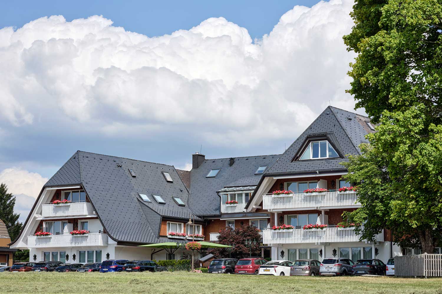 Ein mehrstöckiges Hotel mit Schieferdach und weißen Balkonen, die mit roten Blumen geschmückt sind, davor geparkte Autos und ein blauer Himmel mit flauschigen weißen Wolken.